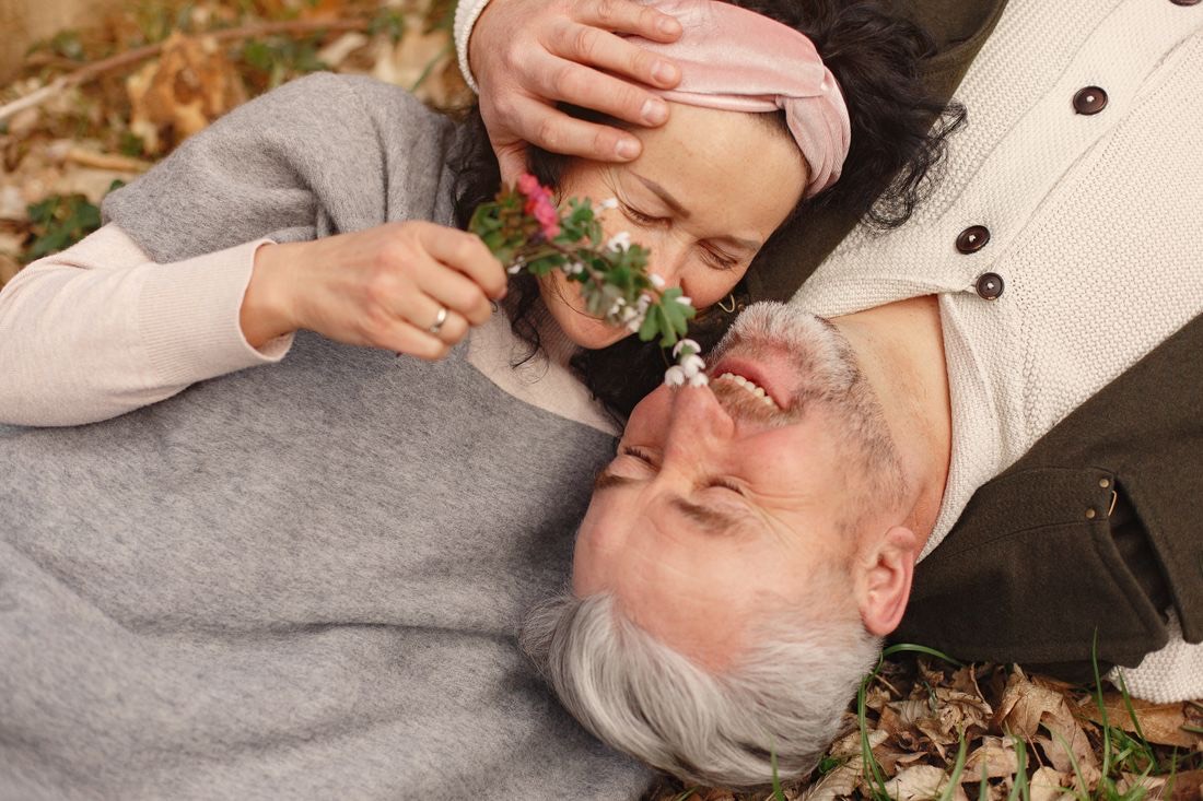 A silver-haired man laughing in the autumn leaves as his wife holds wildflowers to his nose