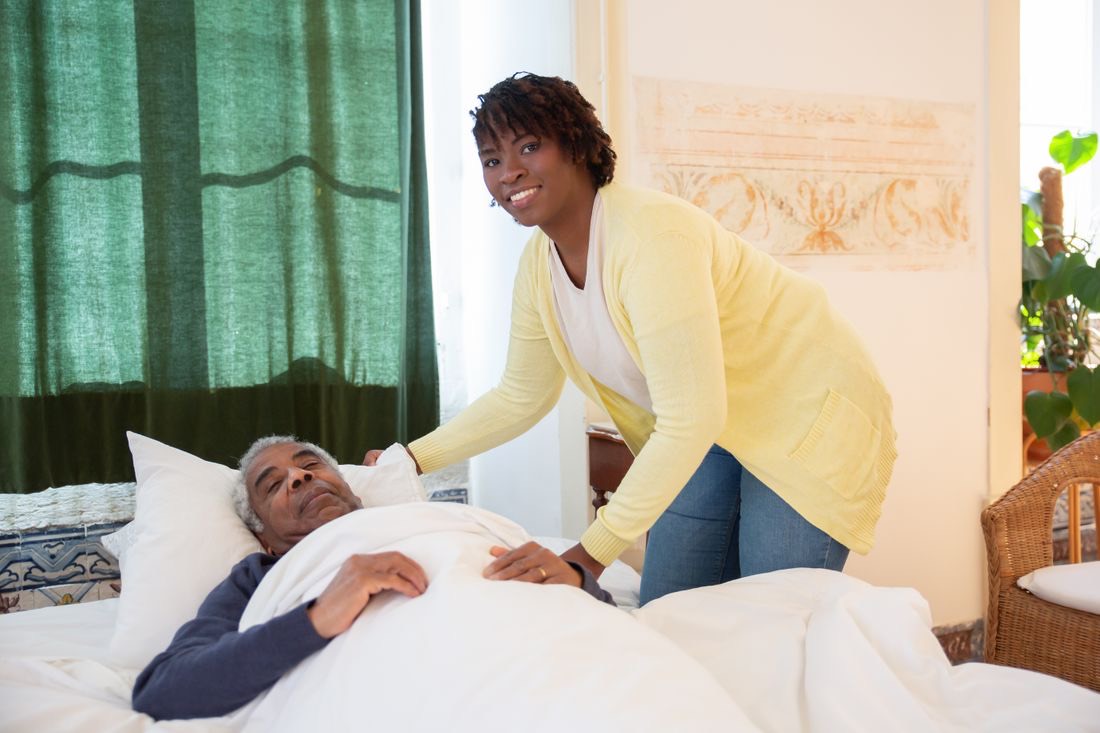 A caregiver warmly tucking in an older man, both smiling
