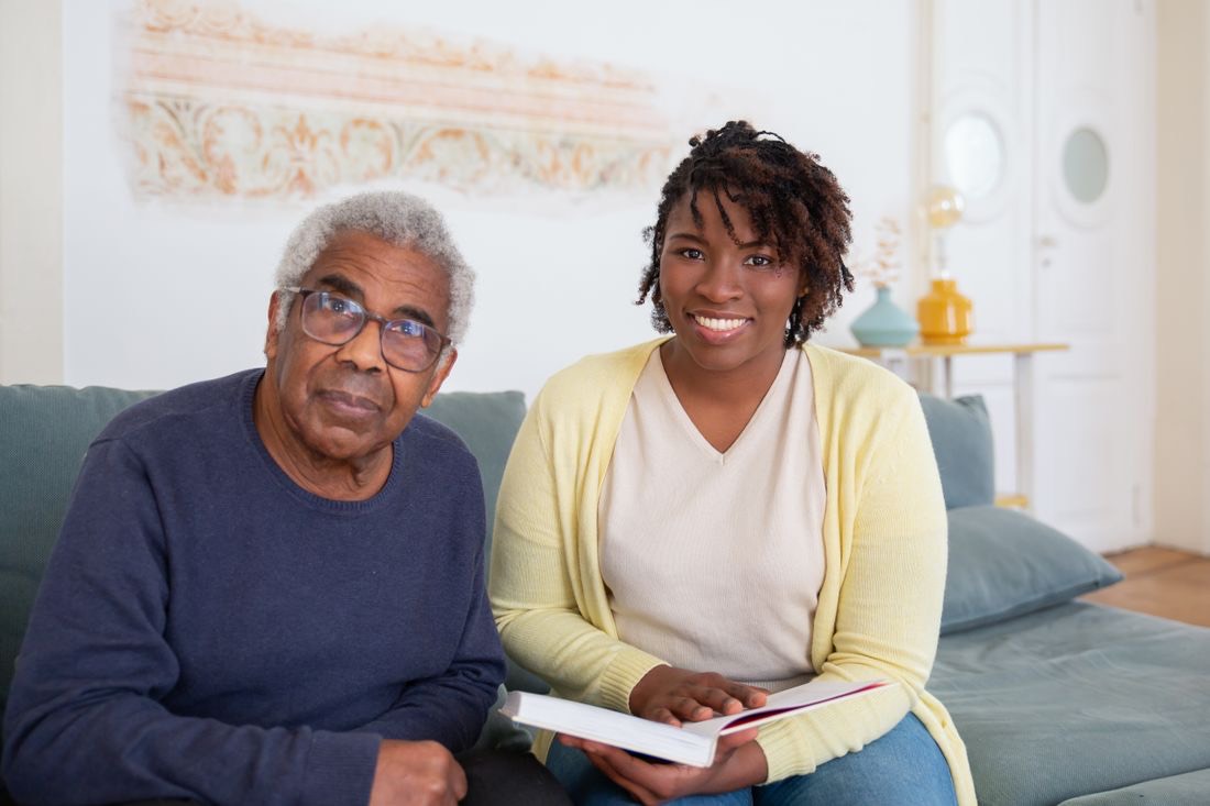 A caregiver sitting with an older man on his couch, reading together